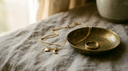 Solid yellow-gold fine jewelry — a cable chain, gold charms and a gold band ring in a brass dish on warm linen