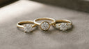 Three solid yellow-gold engagement ring settings — prong solitaire, bezel and three-stone — with colourless lab-grown diamonds on greige suede