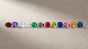 Twelve birthstones in a row on greige linen — garnet, amethyst, aquamarine, diamond, emerald, alexandrite, ruby, peridot, sapphire, pink tourmaline, citrine and topaz