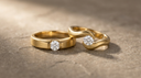 Two solid yellow-gold lab-grown diamond pinky rings on warm greige stone — an architectural east-west band and a sculptural wave band, each set with a single diamond